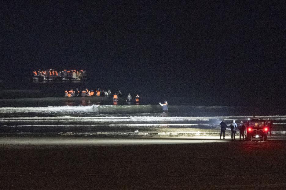 Migrants attempt to board an inflatable boat on a beach at night, while police officers stand nearby.