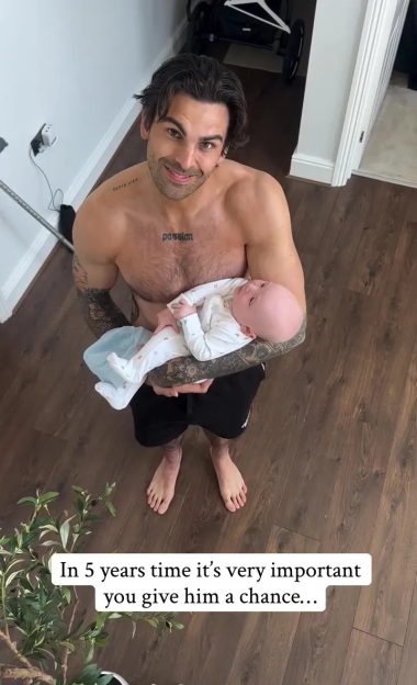 A smiling shirtless man with tattoos holding a baby.