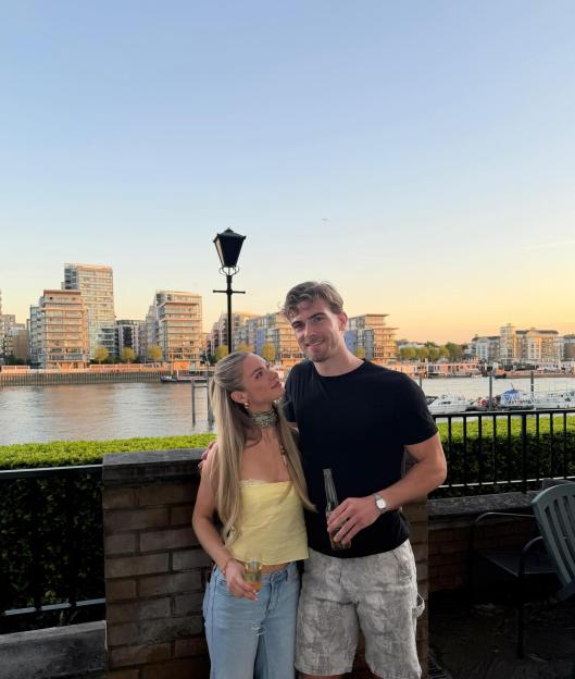 Livvy and Joel Evans posing together by a river with buildings and boats in the background.