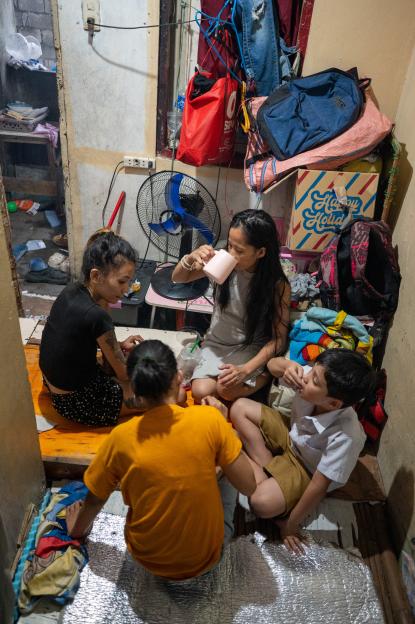 A family of four, David Jurado, his mother Abigail Ocampo Jurado, and her partner Phama Mae Pablo Faurado, eat dinner in a cramped room with a fan and backpacks.