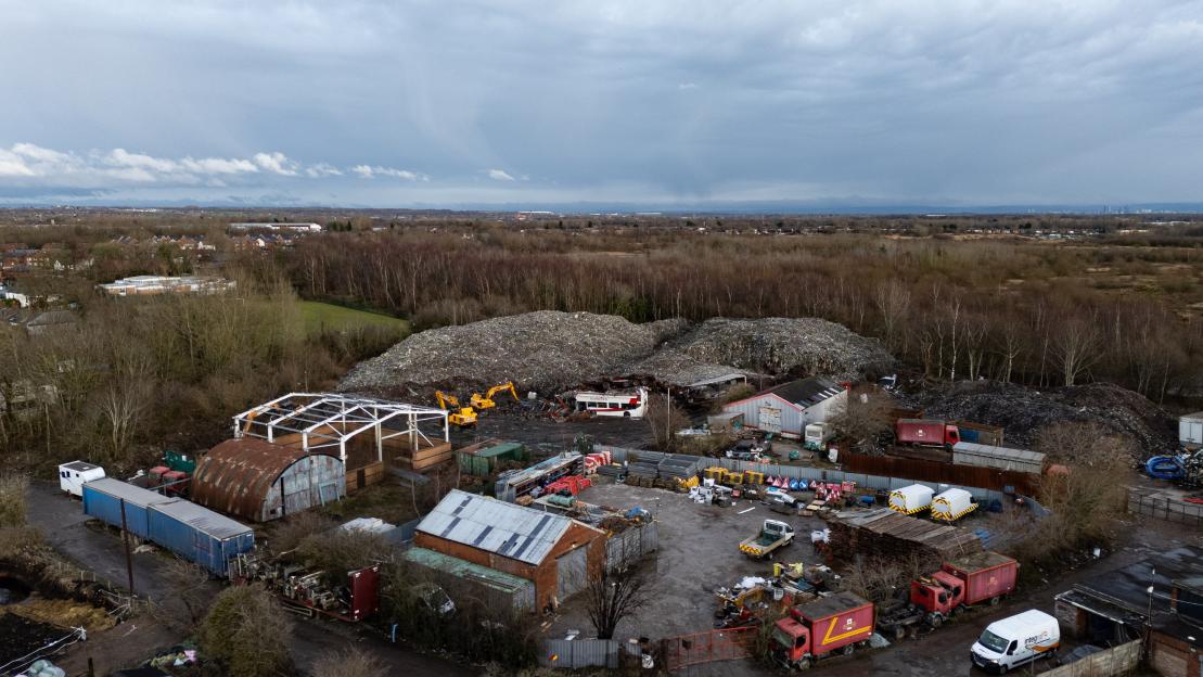 Aerial view of an illegal waste dump, with large piles of refuse, industrial buildings, trucks, and equipment, surrounded by bare trees and distant housing under a cloudy sky.