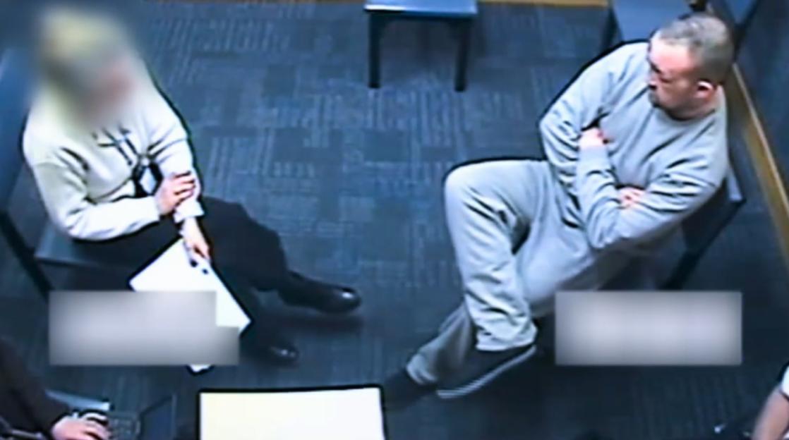 A man in a light grey tracksuit laughs during a police interview as a blurred detective watches.