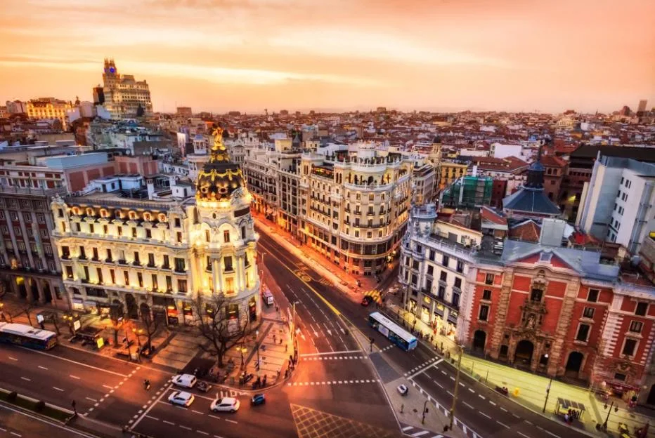 Aerial view of Gran Via in Madrid at dusk, with buildings illuminated and a vibrant orange sky.