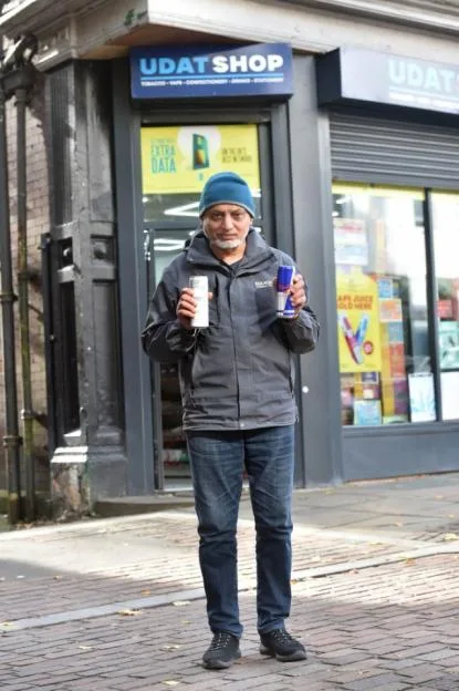 A shopkeeper wearing a blue beanie and a grey jacket holds two drink cans outside his Udat Shop.