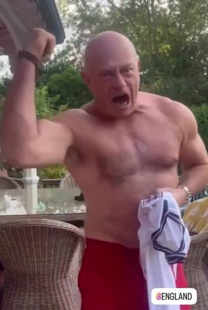 Topless Ross Kemp cheers on England, flexing his arm with an open mouth, wearing red shorts and holding a white shirt.