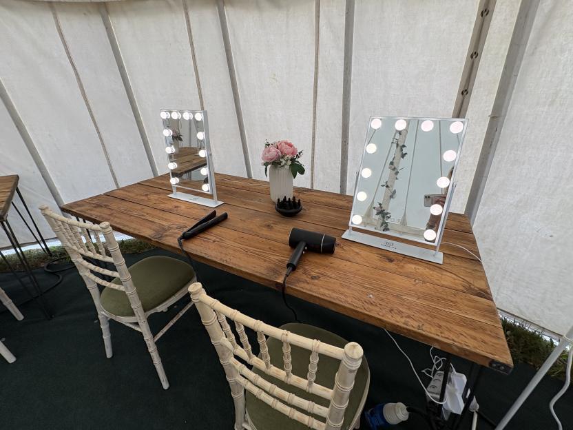 Two vanity stations with lighted mirrors, hair straighteners, and a hairdryer on a wooden table, inside a white canvas tent.