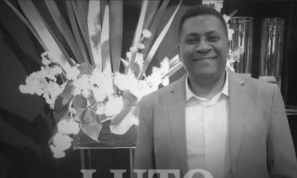 Black and white photo of a smiling man in a suit jacket and collared shirt, standing in front of a floral arrangement and a "LUTO" sign.