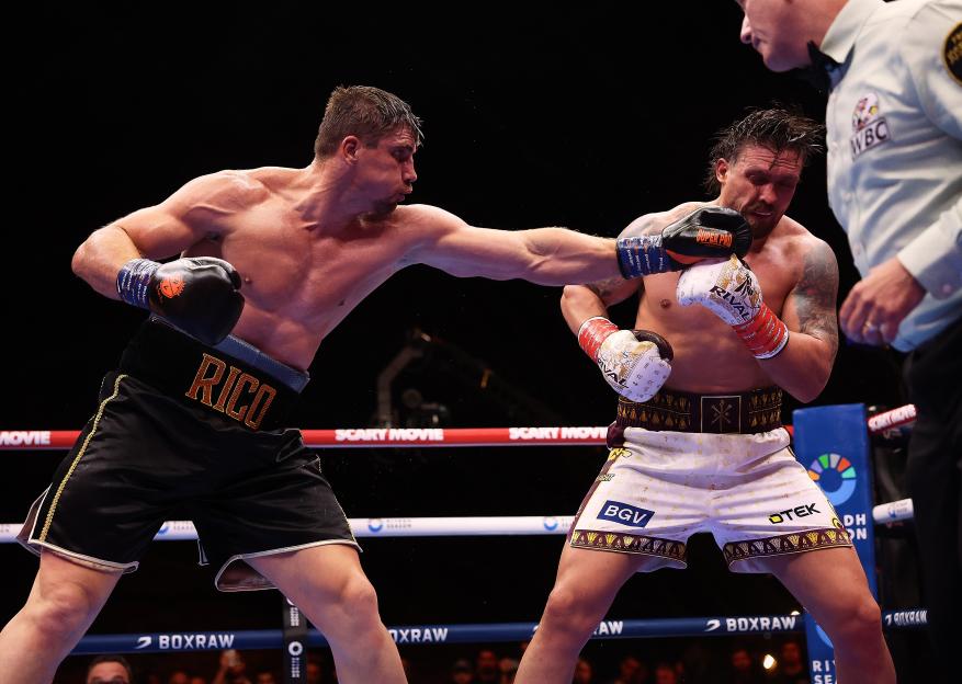 A boxer wearing black shorts with "RICO" in gold lettering punches an opponent in white shorts.