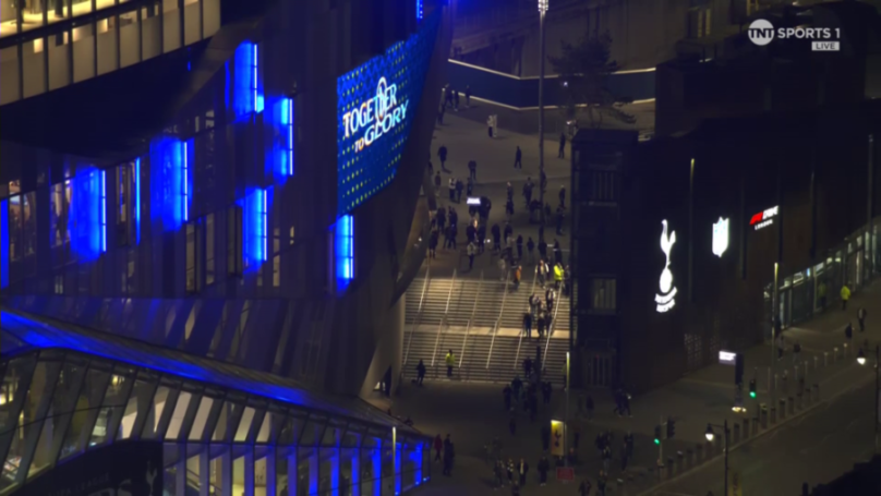 A night-time view of Tottenham Hotspur Stadium, with a large screen displaying "TOGETHER TO GLORY" and the Spurs logo on a nearby building.