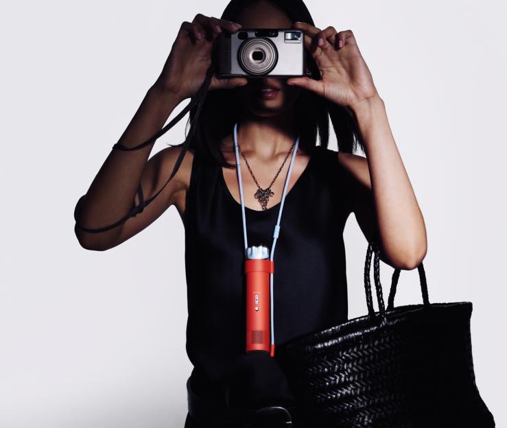 A woman holding a camera to her face, wearing a black top, with a Dyson HushJet Mini fan and a black woven handbag.