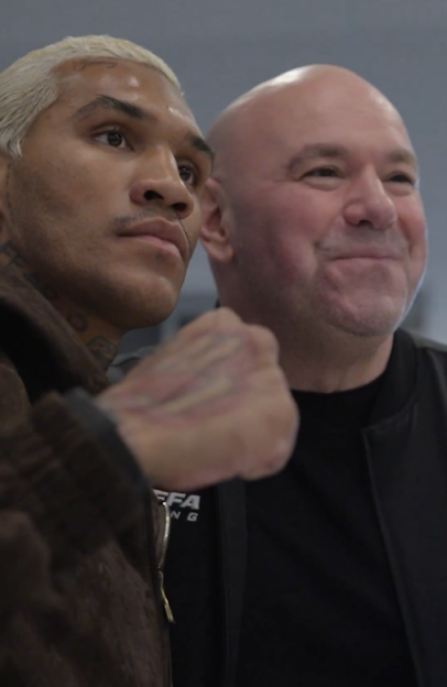A man with blonde hair and a beard, wearing a brown jacket, stands next to a smiling bald man in a black jacket.