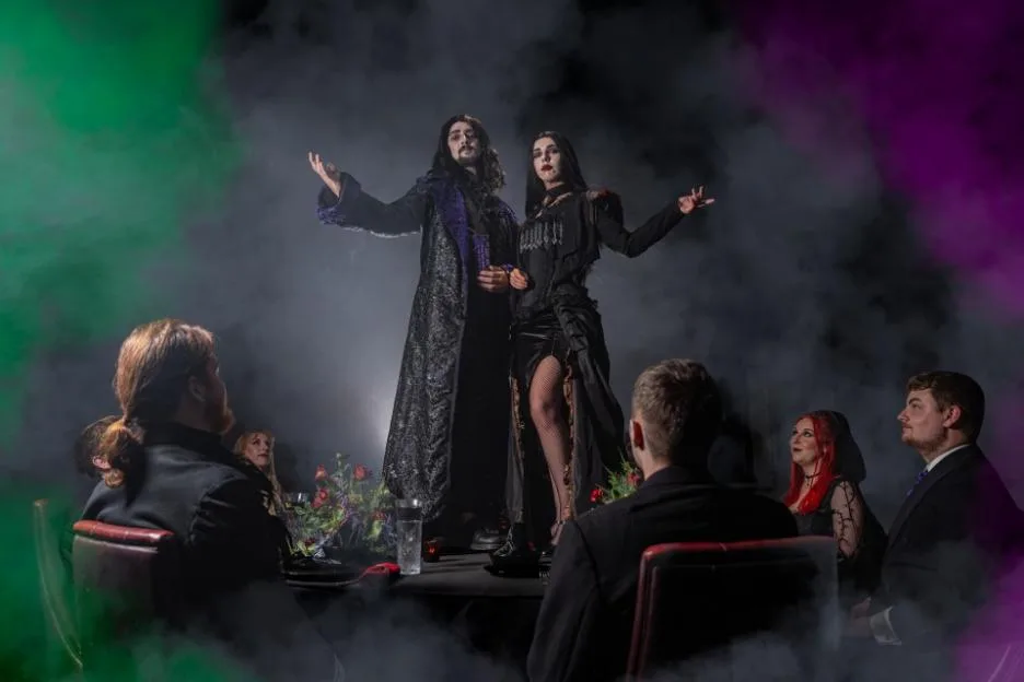 A man and a woman in vampire-like attire stand at the head of a dinner table, surrounded by smoke and seated guests.