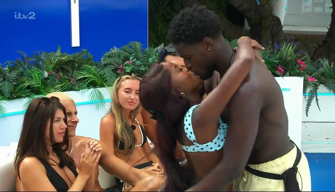 A couple kissing in a reality TV show while a woman in a bikini looks on with a side-eye.
