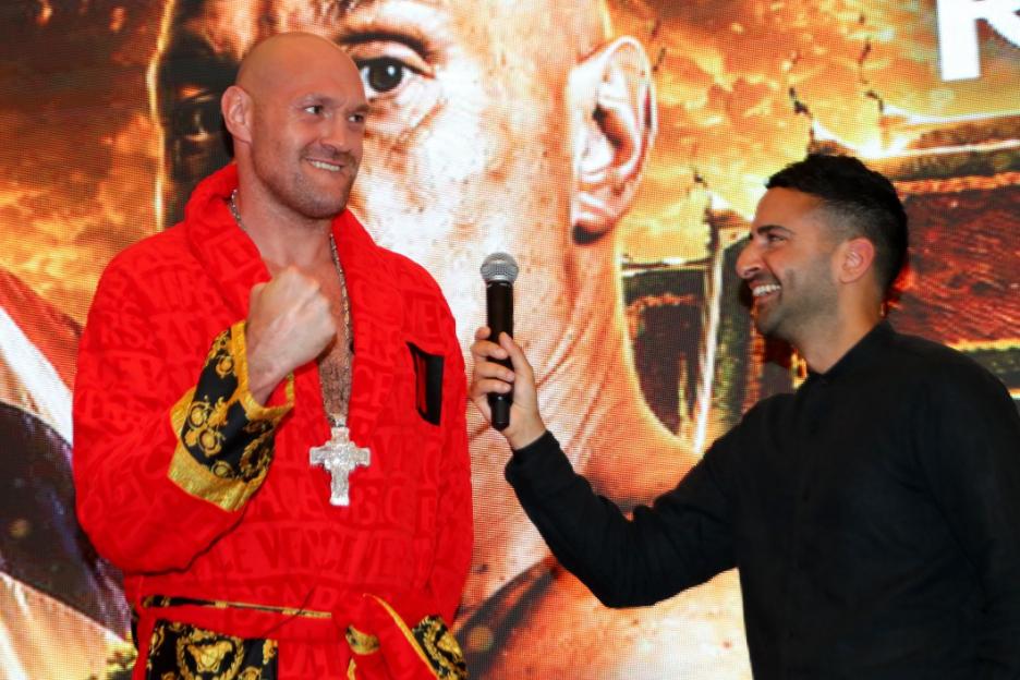 Tyson Fury wearing a red robe and a silver cross necklace, being interviewed by a man holding a microphone.