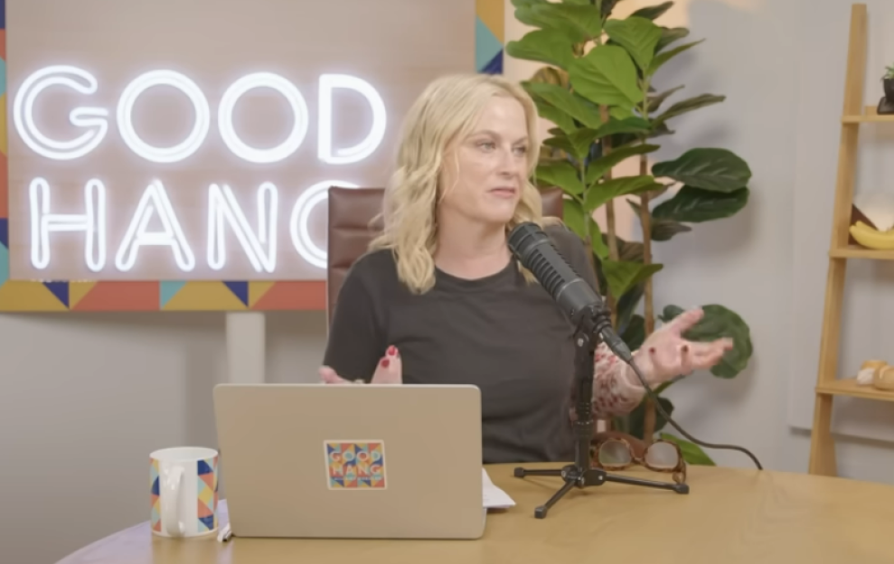 Woman with blonde hair speaking into a microphone with a laptop and a "Good Hang" neon sign in the background.