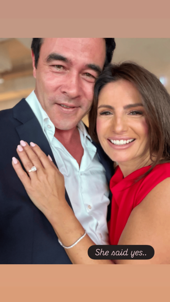 A man in a suit and a woman in a red top, with a diamond ring visible on her hand, smile at the camera, with the text "She said yes.." at the bottom right.