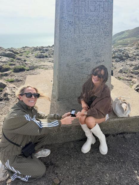 Fran Parman proposes to Molly McCann-Pearson, who is seated on a stone base, holding out an engagement ring box.