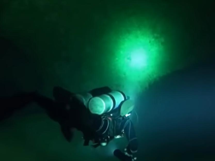 A diver in dark water with a green light illuminating a wall.