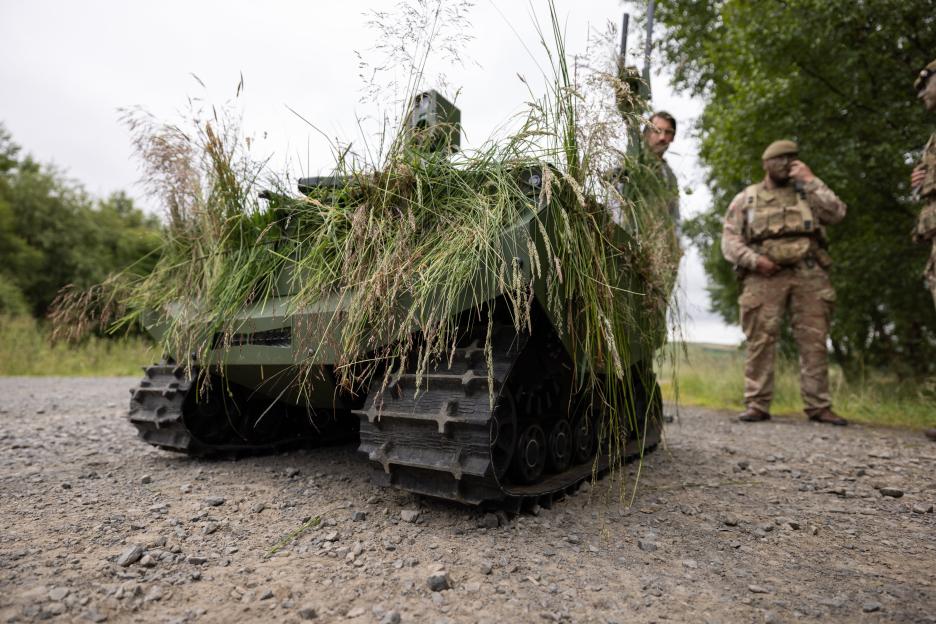 A Gereon uncrewed ground vehicle, camouflaged with tall grass, sits on a gravel path as soldiers stand in the background.