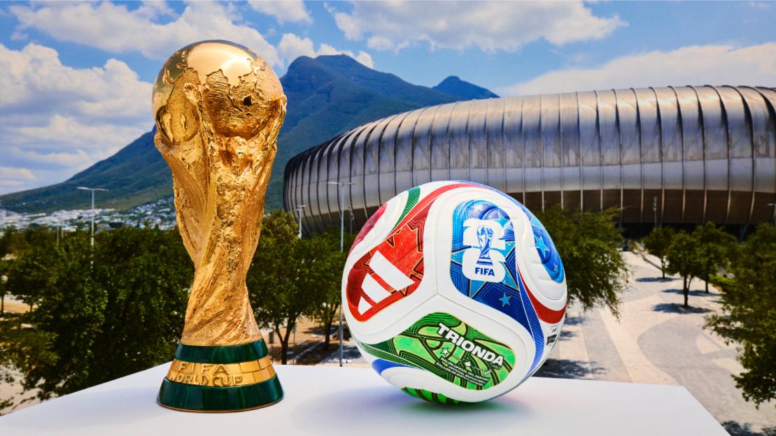 The FIFA World Cup trophy next to the red, green, and blue Trionda soccer ball with a stadium and mountains in the background.