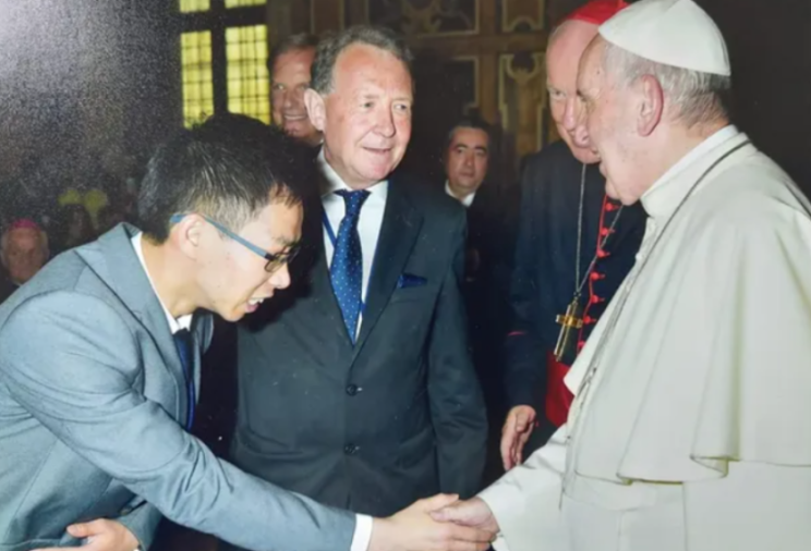 A man in glasses and a suit shakes hands with the Pope, while another man in a suit and tie smiles in the background.