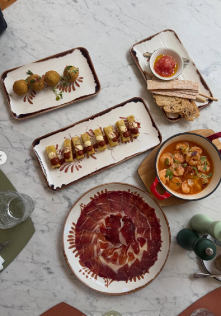 A variety of tapas dishes, including croquettes, sausage rolls, sliced ham, sautéed shrimp, and bread with tomato spread.