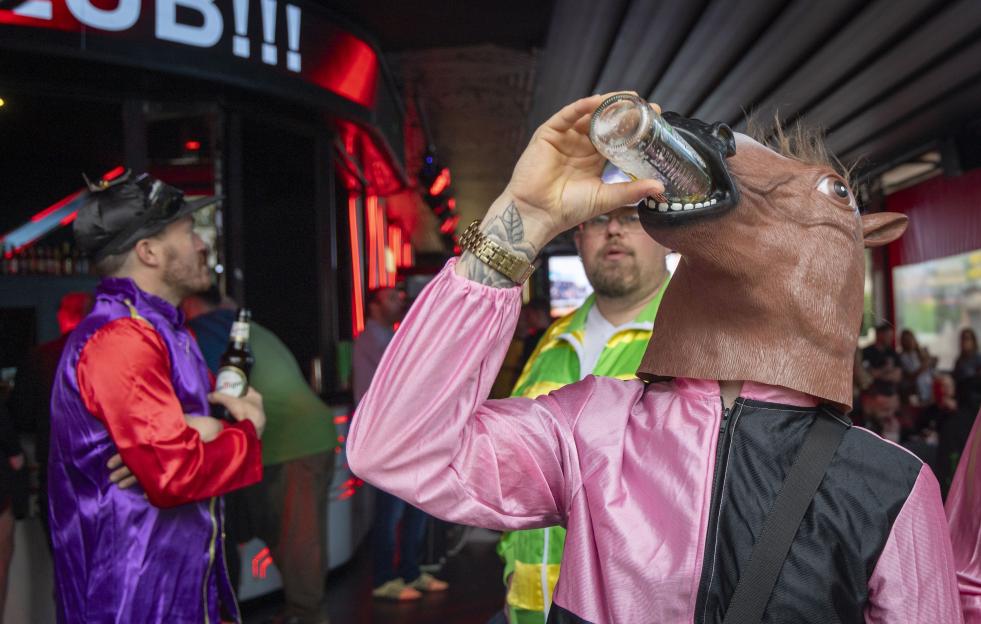Man in horse mask drinking a bottle of beer with Cheltenham race fans.