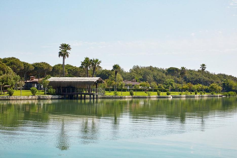 Gloria Golf Resort, showing a thatch-roofed structure over water next to a lush green shoreline with palm trees and a hotel building.