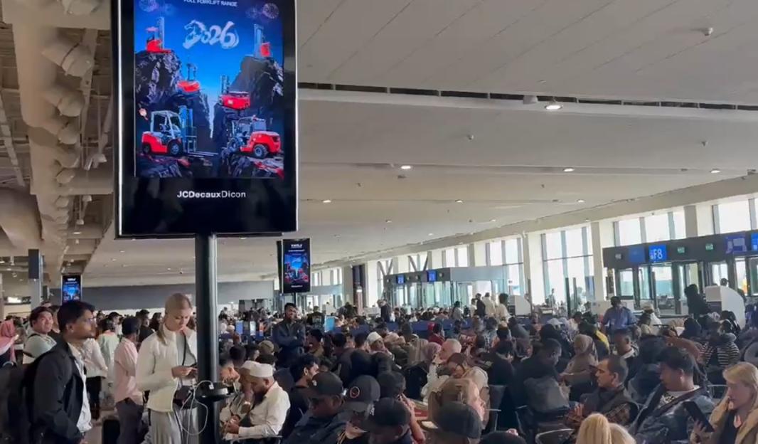 A large crowd of people waiting at an airport terminal, with an advertisement for JCB forklifts visible on a large screen.