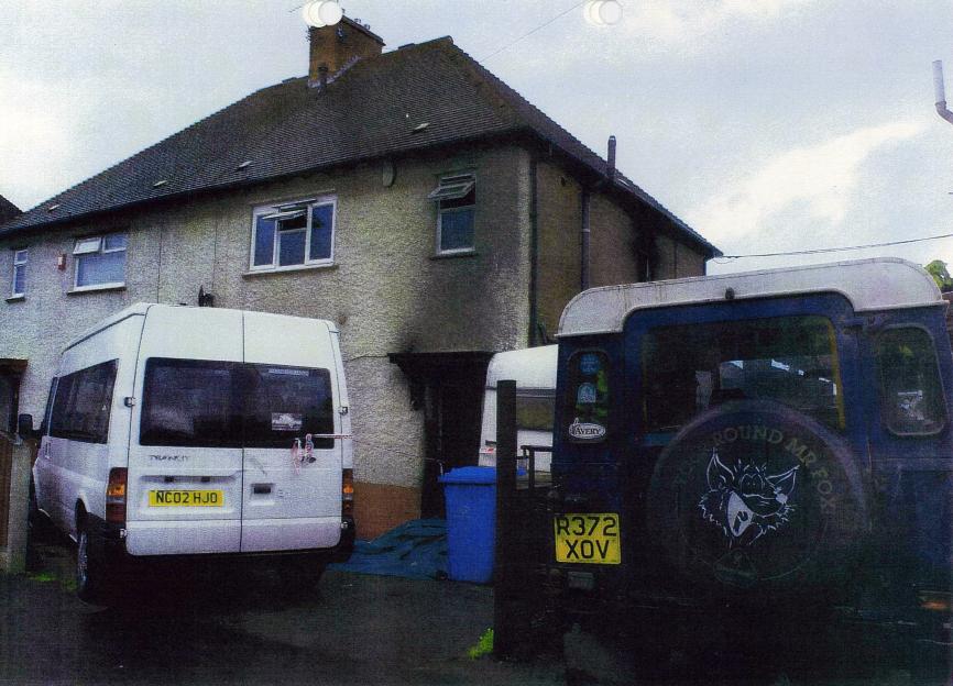 A two-story house with a blackened door and charring around a second-story window, with a white van parked in front of it and a dark blue vehicle with a spare tire on the back parked to the right.