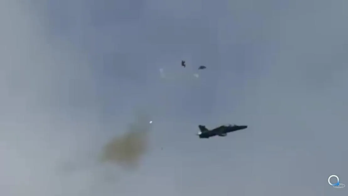 A fighter jet flying with a cloud of smoke and two parachutes visible in the sky.