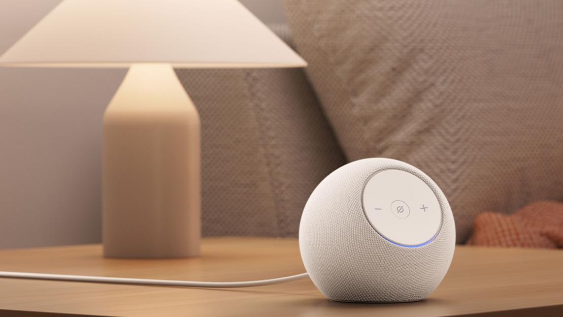 A white fabric-covered smart speaker with a blue light at its base on a wooden surface next to a lamp.