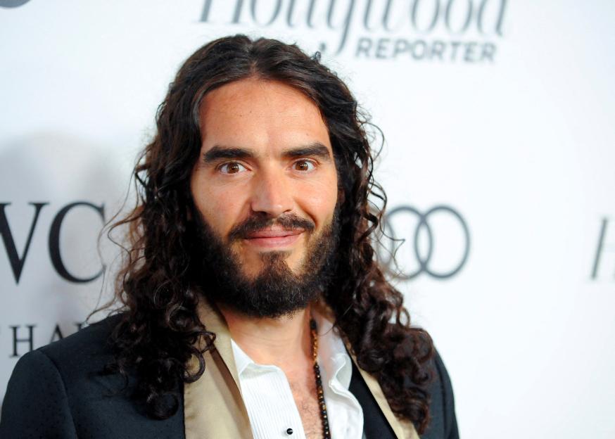 British actor and comedian Russell Brand with dark curly hair and a full beard, wearing an open white shirt and dark jacket.