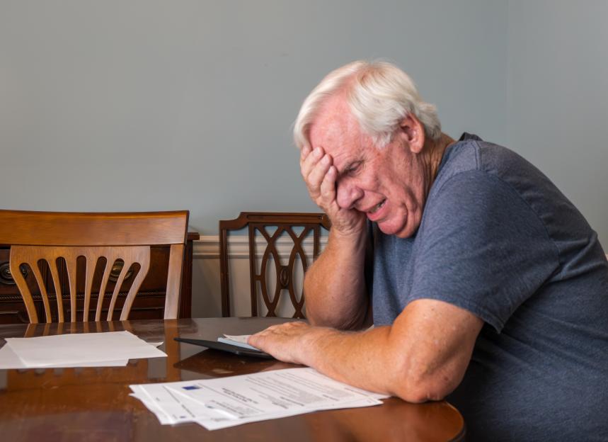 Stressed senior man with white hair, wearing a gray t-shirt, sits at a table and clutches his forehead while looking at bills.
