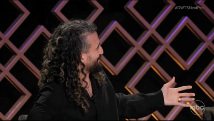 A man with long curly hair and a beard, wearing a black shirt, smiles while extending his left hand.