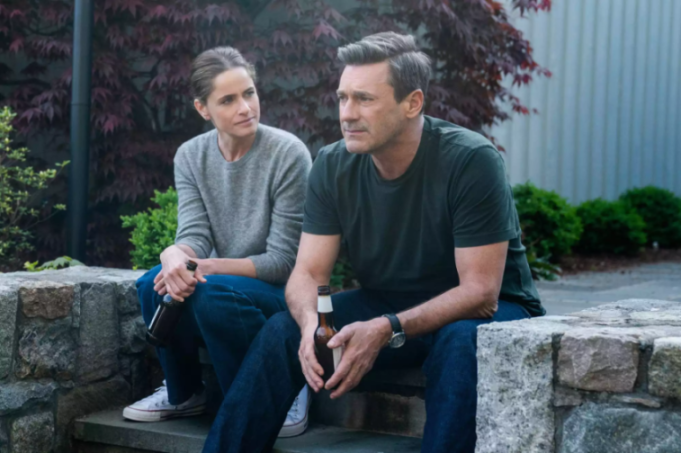 Jon Hamm and Amanda Peet sitting on a stone wall, holding beer bottles.