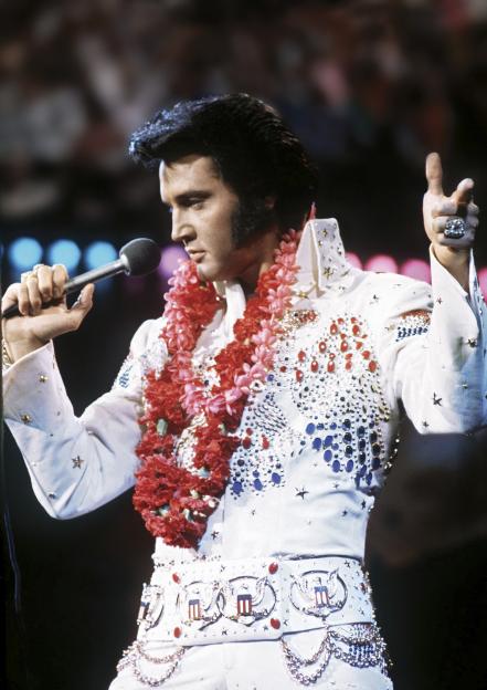 Elvis Presley singing into a microphone, wearing a white jumpsuit decorated with jewels and a red floral lei, during his Aloha from Hawaii NBC special.