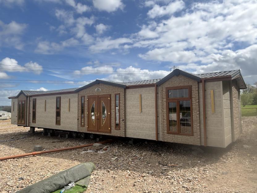 A luxury mobile home, built to resemble a permanent house with stone siding and a shingled roof, sits on a gravel lot under a partly cloudy sky.