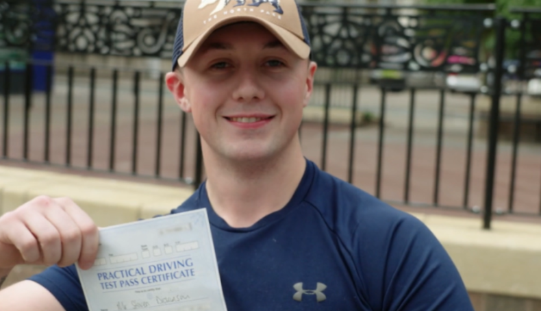 A young man in a baseball cap smiles while holding up a "Practical Driving Test Pass Certificate."