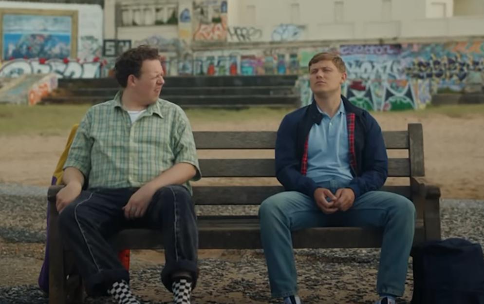 Two men sitting on a park bench, with buildings and graffiti in the background, from the show Big Boys.