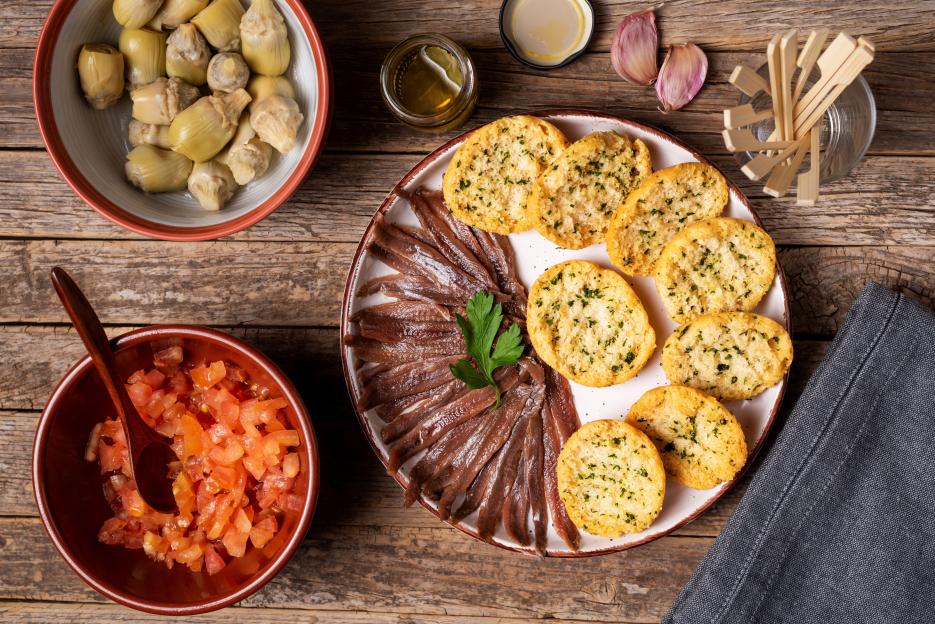 Cantabrian anchovies, toast with parsley, chopped tomato, artichokes, garlic, and olive oil, top view.