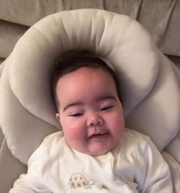 A baby with dark hair and rosy cheeks wearing a white outfit with turtle illustrations, sitting in a padded baby seat.