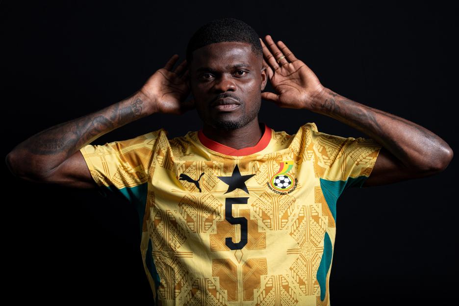 Thomas Partey posing in a yellow and green Ghana football jersey with a black background.