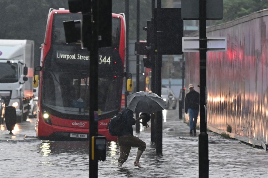 A person holding an umbrella and walking barefoot through a flooded street as a red bus passes by in London.