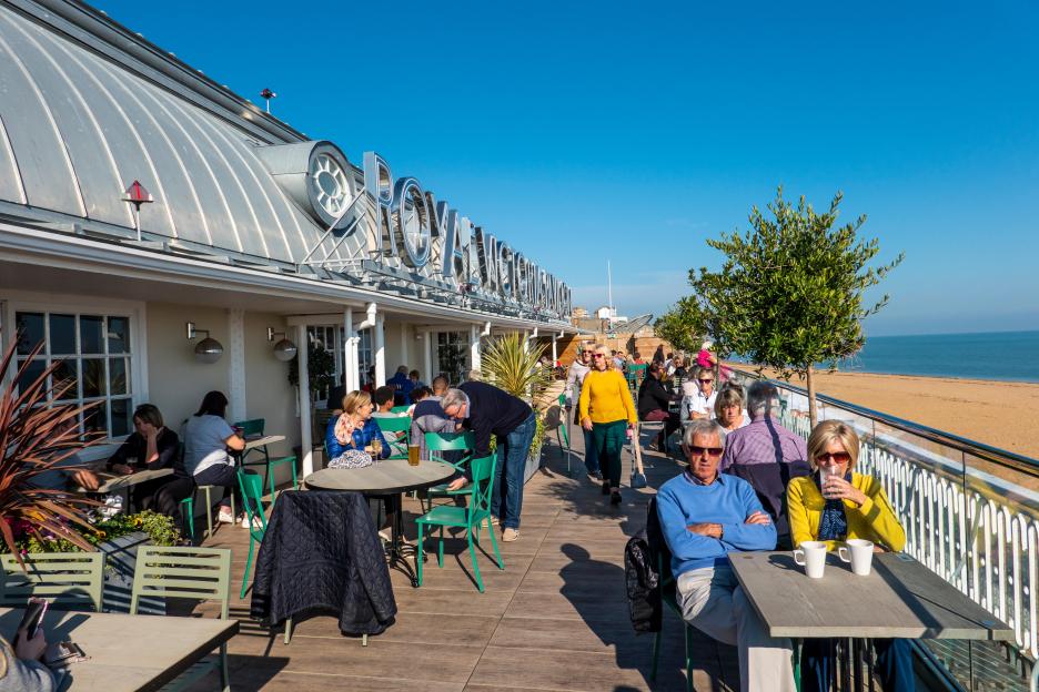 People relaxing at an outdoor café on a pier overlooking a sandy beach and the sea.