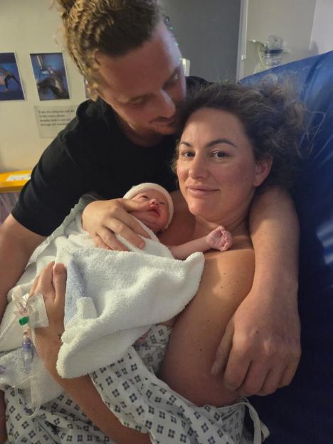A mother holds her newborn baby, wrapped in a white blanket and wearing a pink hat, while her partner embraces her from behind, in a hospital room.