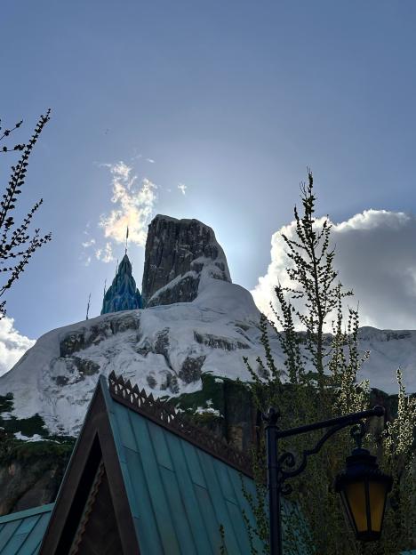 Snow-capped mountains with an ice castle on the left, partially obscured by sun and clouds, with a dark roof and street lamp in the foreground.