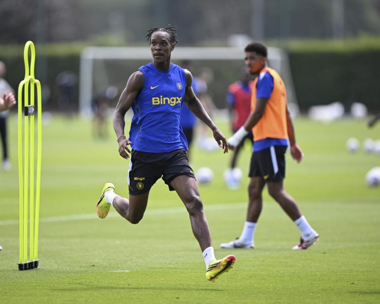 A Chelsea FC player in a blue training vest and black shorts runs on a green field.
