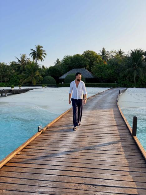 A man walks on a wooden pier over turquoise water with a sandy beach and palm trees in the background.