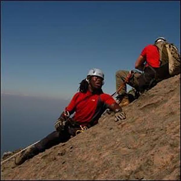 Ade Adepitan rappelling down a rocky slope with another person in the background.